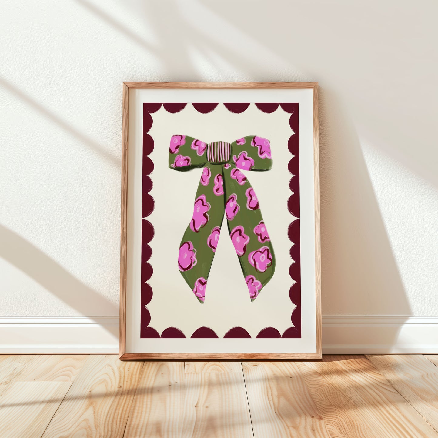 A framed art print depicting a whimsical pink and green floral bow against a scalloped border, displayed on a wall.
