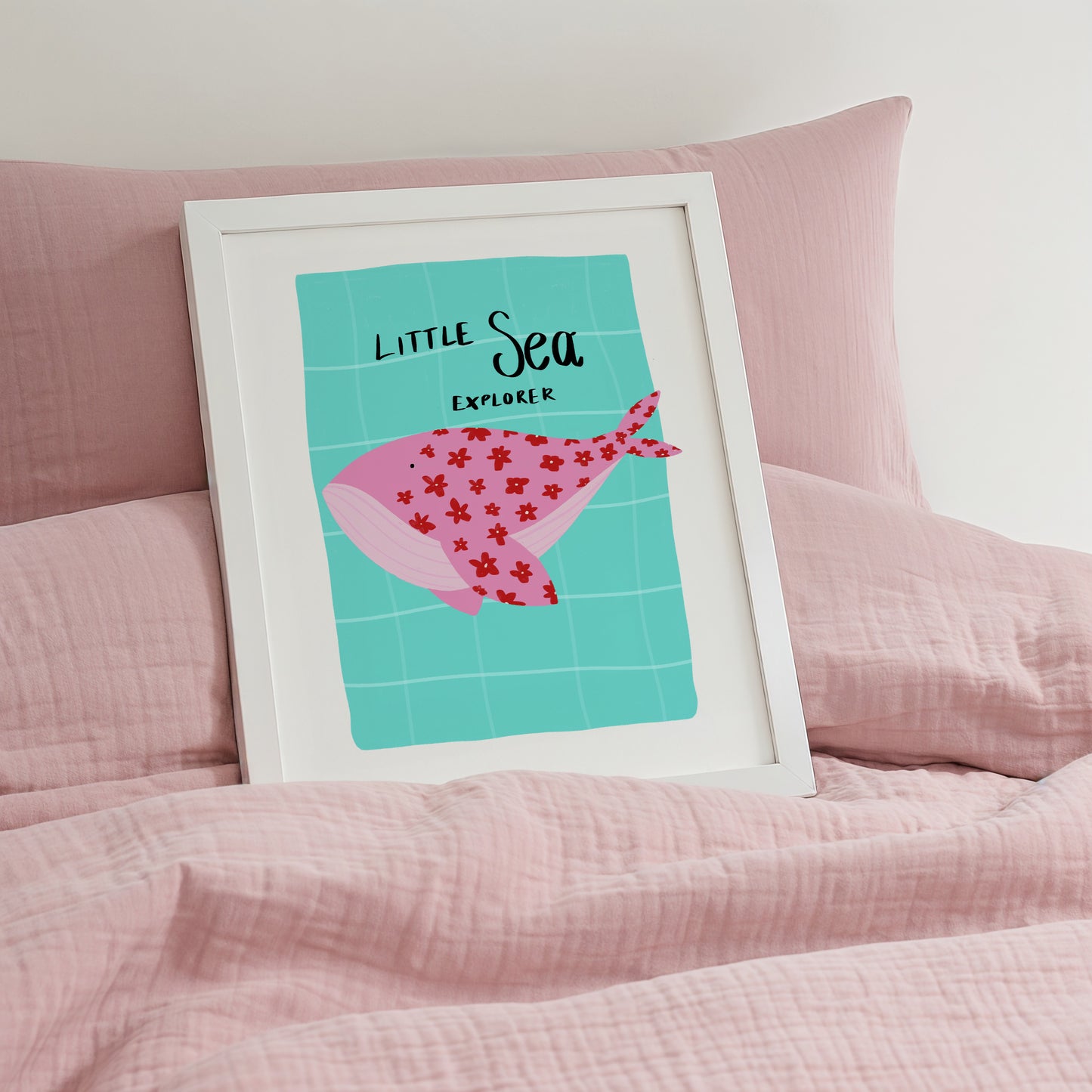 Framed artwork with a pink whale illustration and 'Little Sea Explorer' text on a light blue background, placed on a pink bed.