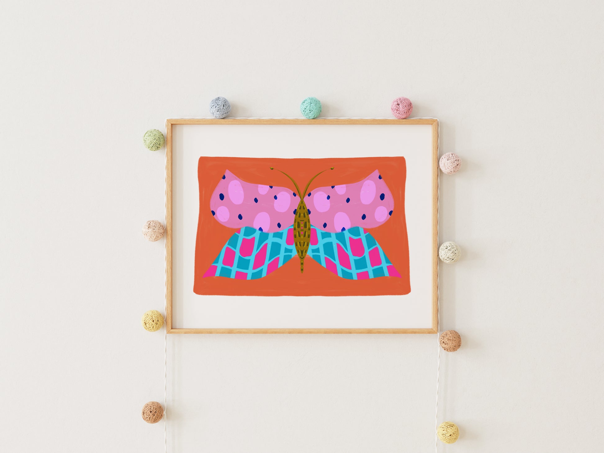 Framed artwork of a pink butterfly on an orange background, hanging on a wall with decorative pom-pom balls.