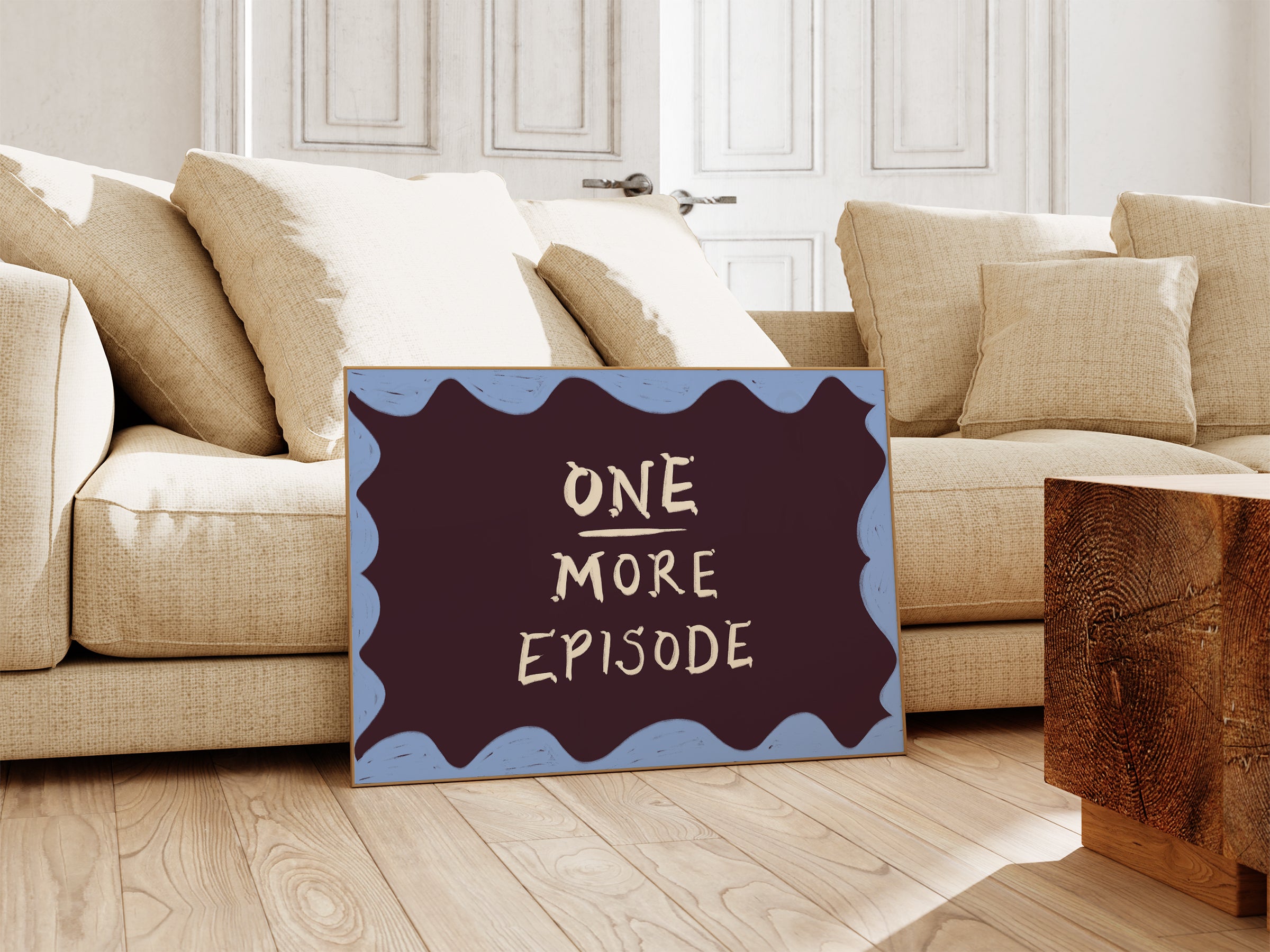 A landscape print leaning against a beige sofa in a bright living room. The print has a dark plum background with wavy light blue borders and the text 'ONE MORE EPISODE' in a light cream handwritten font.