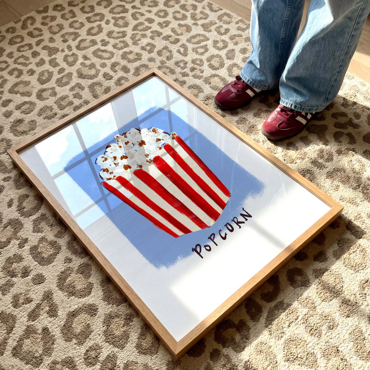 Popcorn art print in a wooden frame featuring a red and cream striped popcorn box on a blue background.