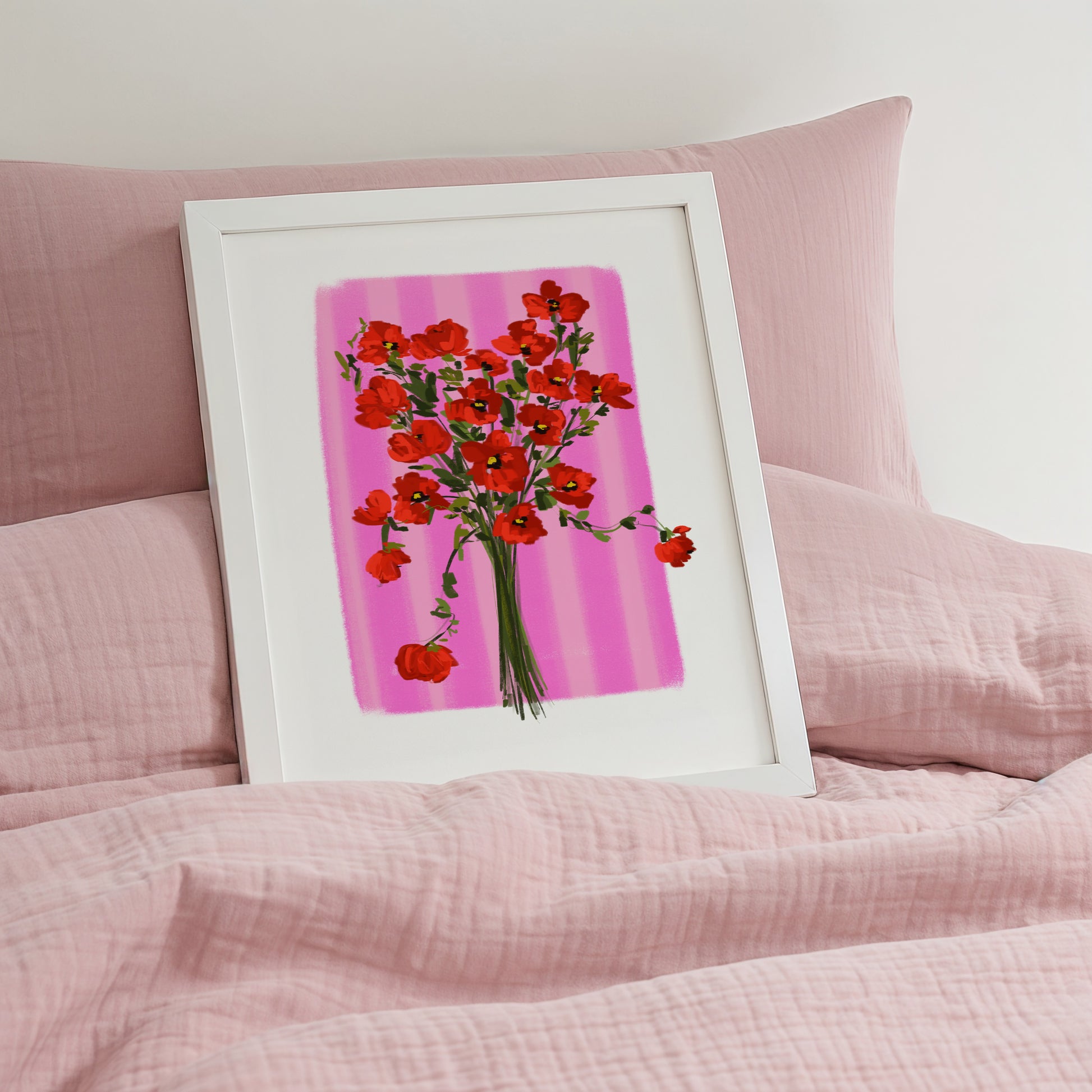 Framed artwork of red flowers on a pink background, placed on a pink bed surface.